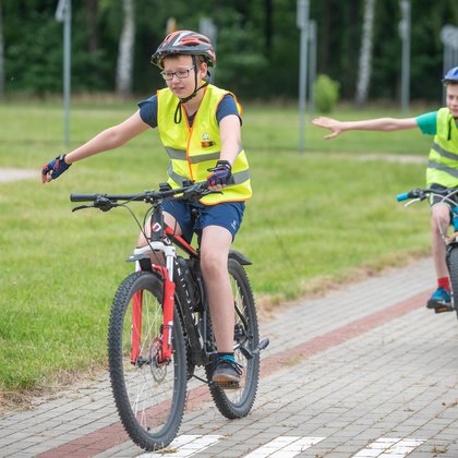 Miasteczko drogowe, dwóch chłopców na rowerach w kaskach i kamizelkach odblaskowych uczy się prawidłowego manewru skrętu w prawo. 